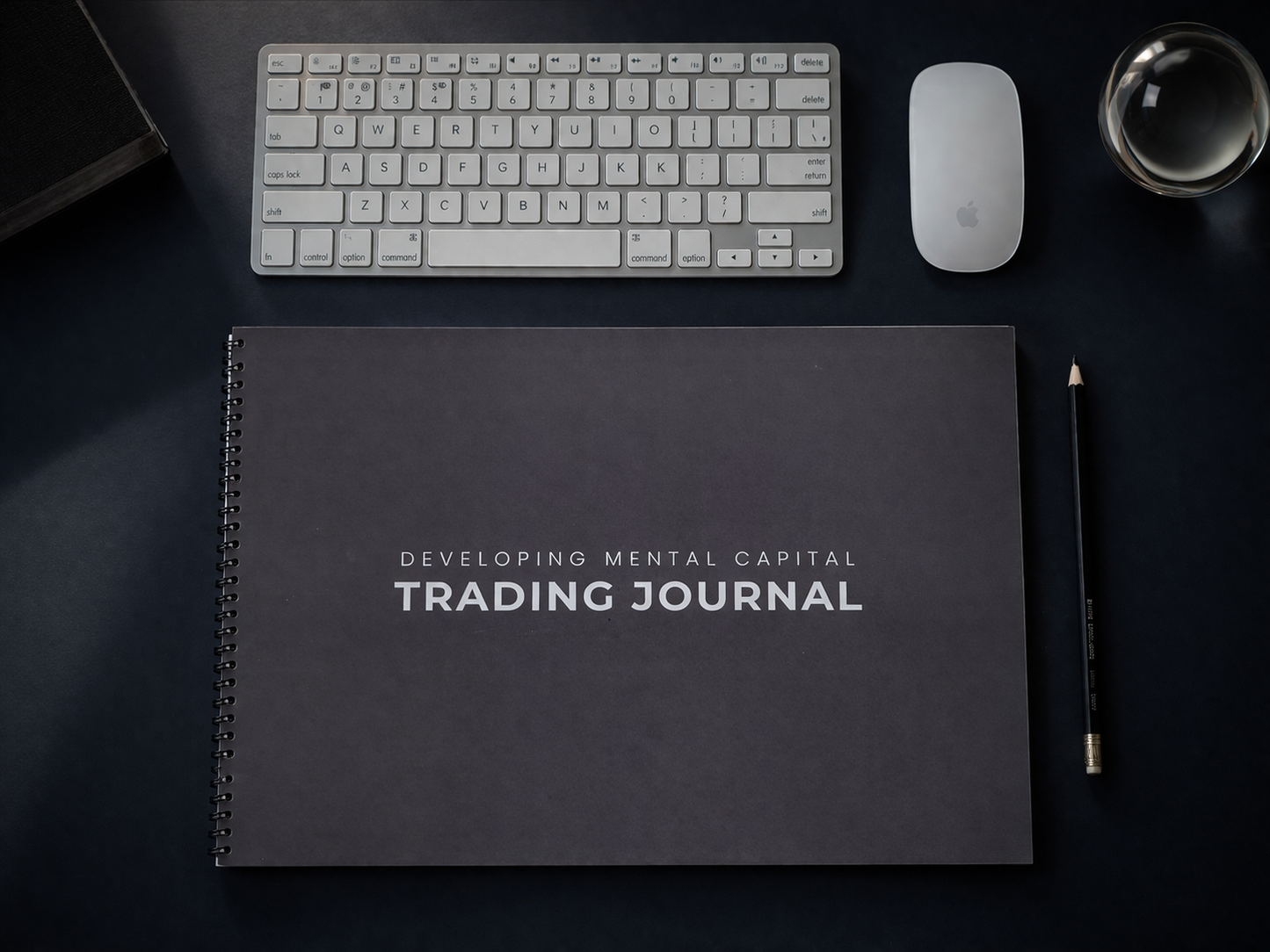 Dark notebook titled 'Developing Mental Capital Trading Journal' on a desk with a keyboard and mouse.